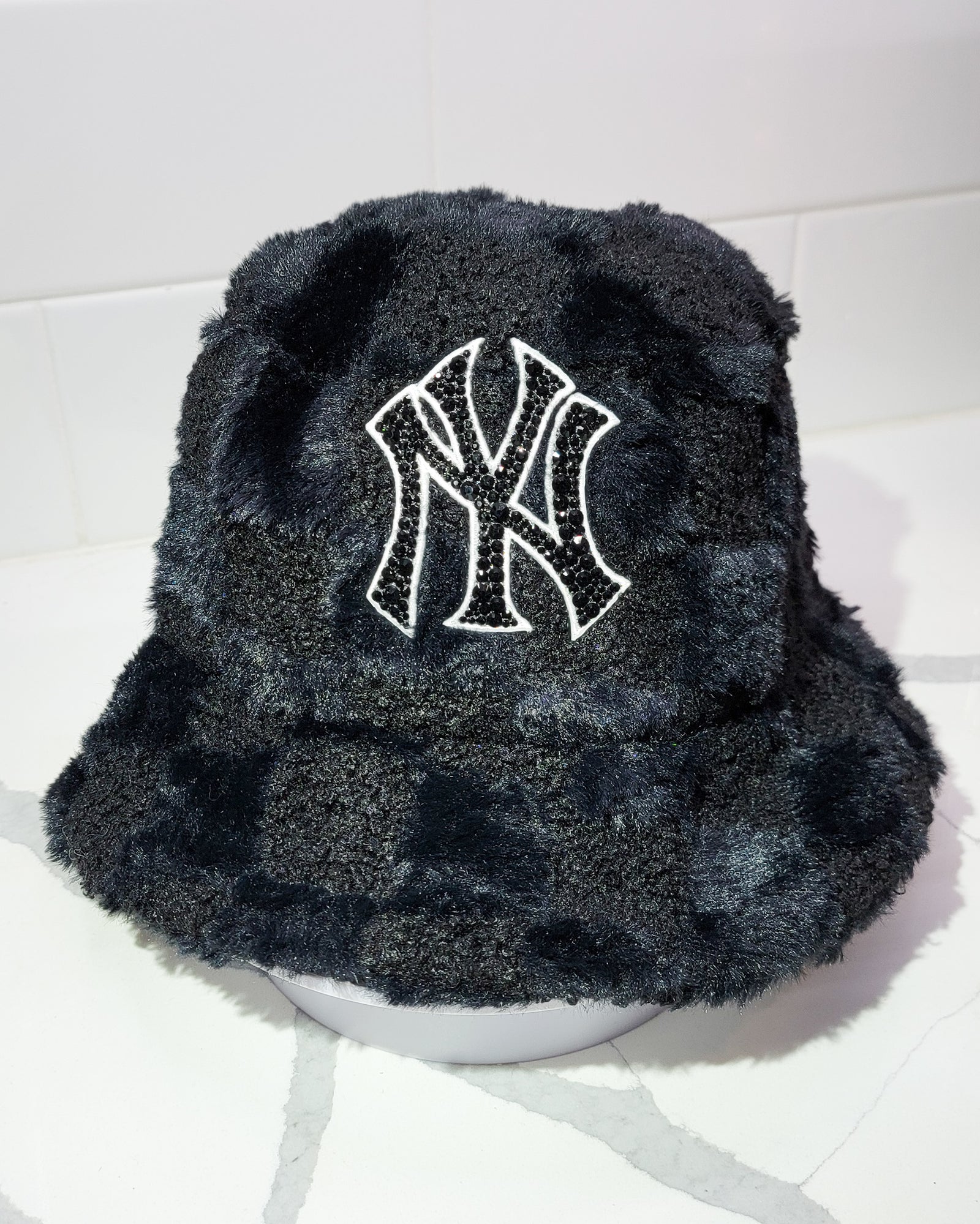 The front of Furry Checkered NYC Bucket Hat, featuring a furry, checkered bucket hat with a prominent logo covered in rhinestones.