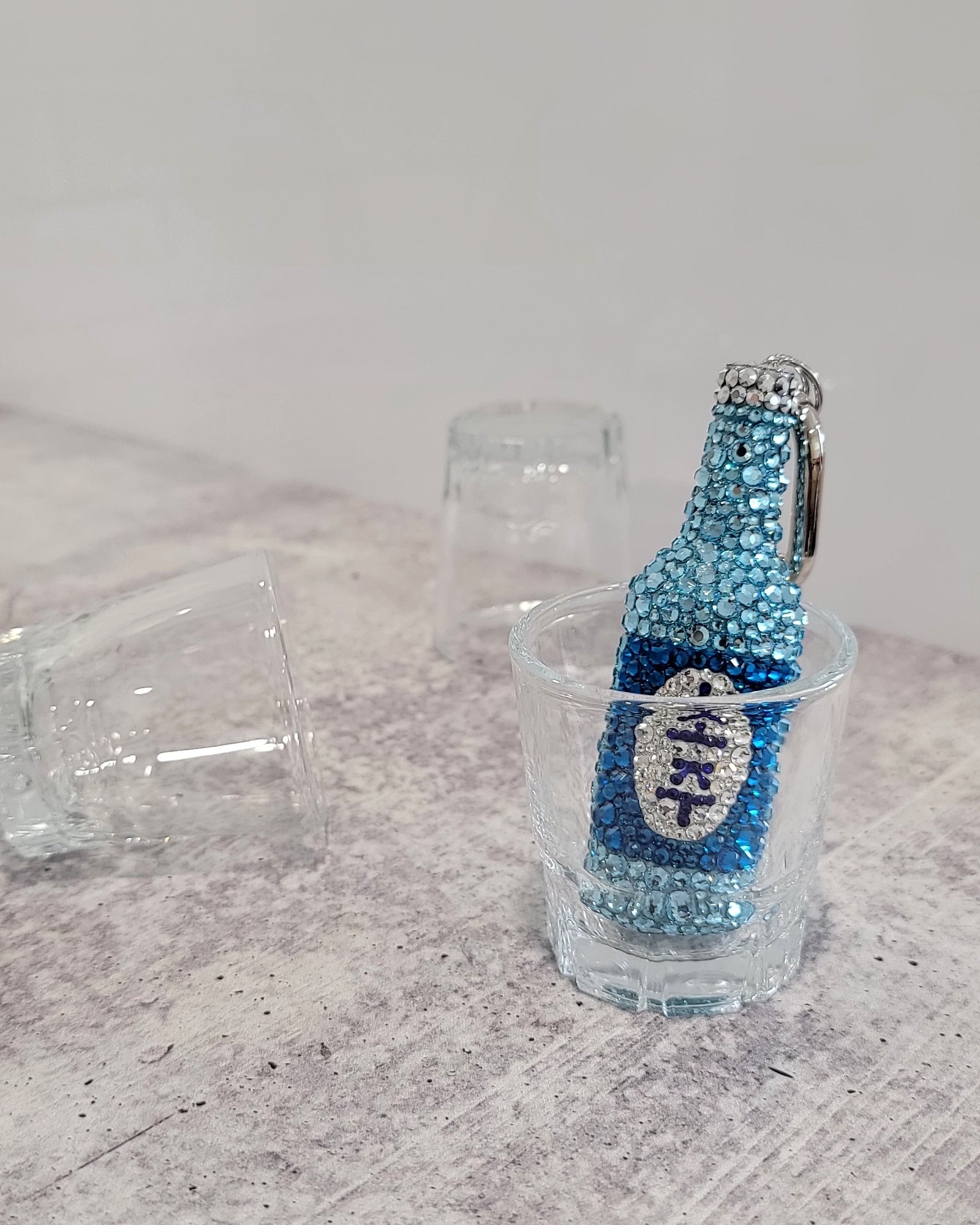 "Fresh" Soju keychain (a blue soju bottle with a blue and white label, featuring silver keychain hardware) sitting inside of a soju cup.