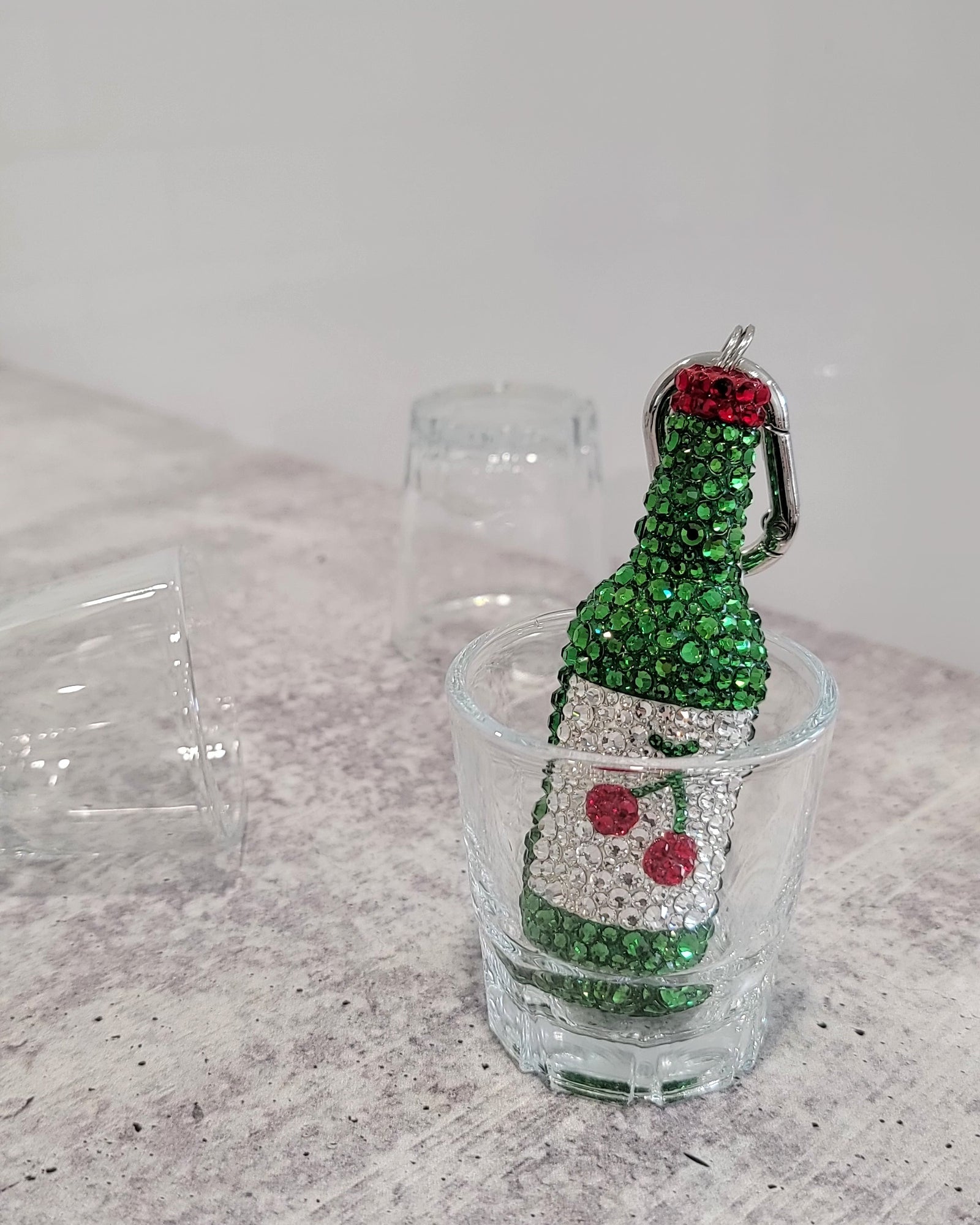 "Cherry" Soju keychain (a green soju bottle with a white label and a small cherry, featuring silver keychain hardware), sitting inside a soju cup.