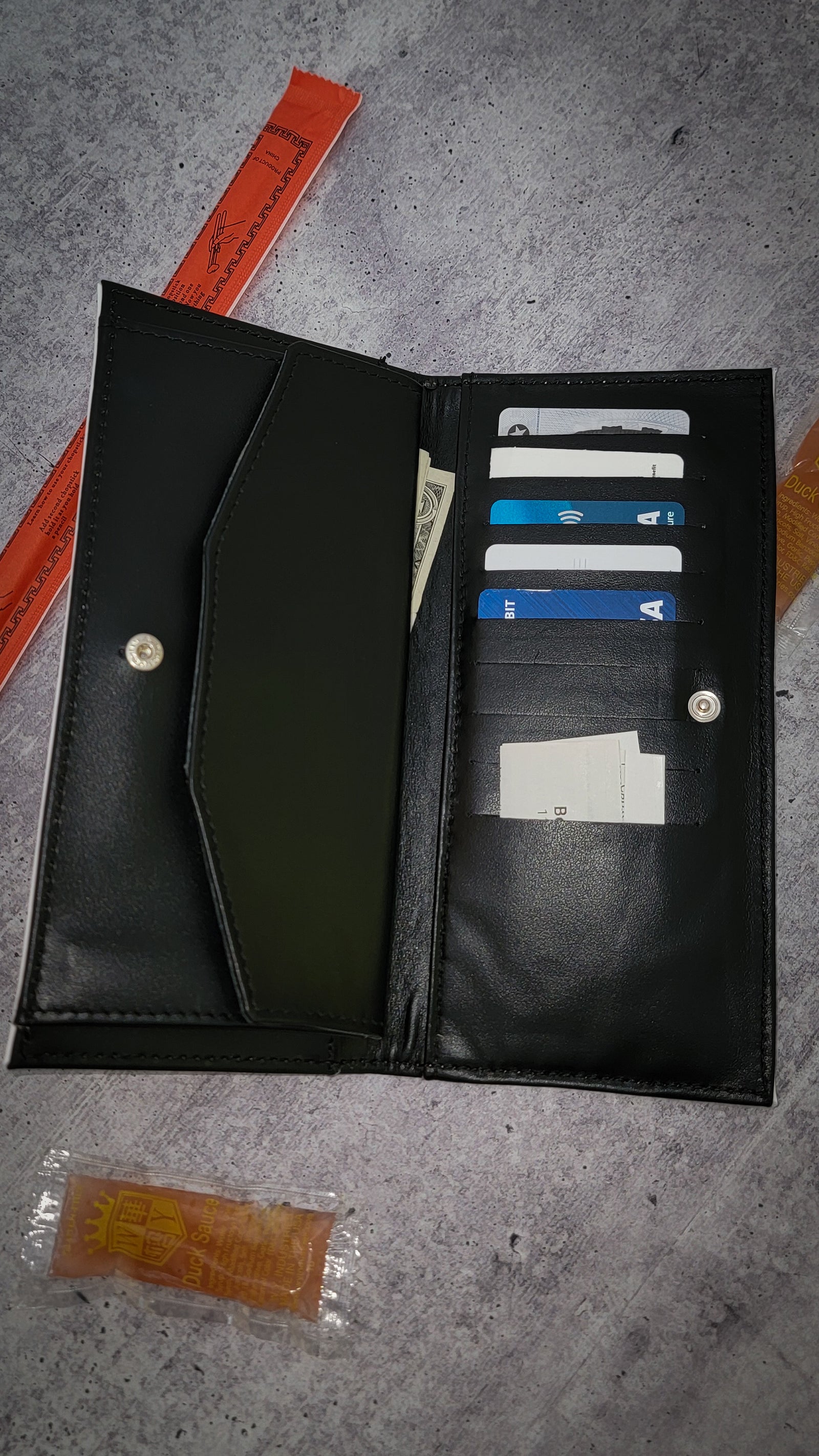The black interior of a leather wallet printed in the style of a Chinese takeout menu, against a gray concrete background, featuring 9 card slots and a hidden pocket for cash.