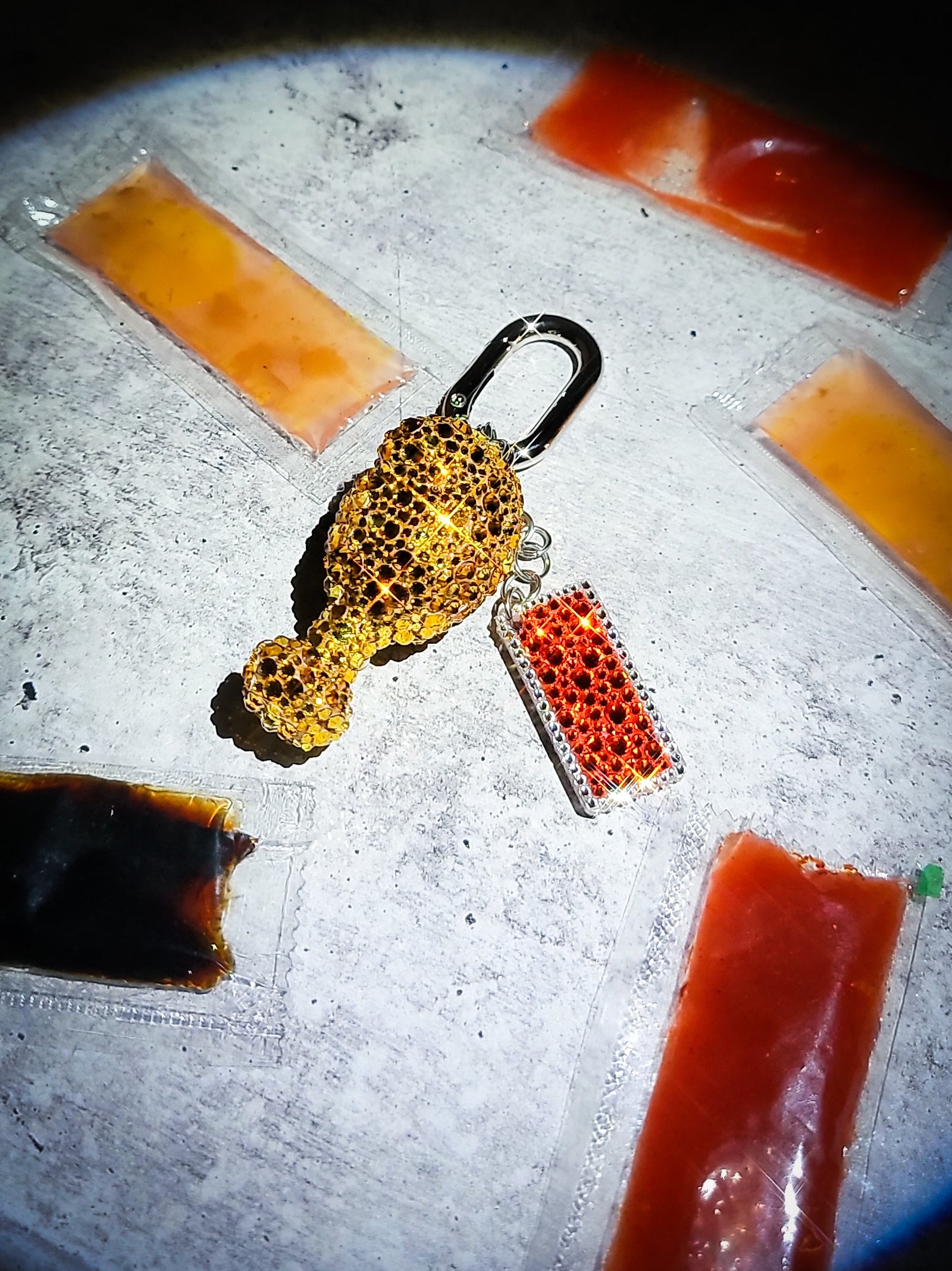 A front view of a chicken drumstick bag charm covered in rhinestones with a tiny hot sauce charm.