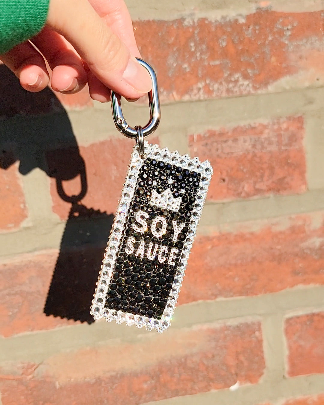 A hand holding a soy sauce bag charm with black background and white text, adorned with white rhinestones, against a brick wall backdrop.