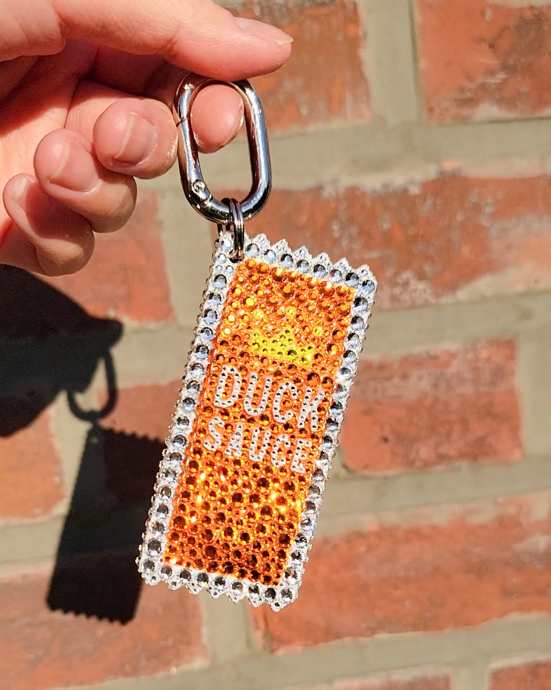 An image of a handmade orange bag charm adorned with Czech Preciosa rhinestones, designed to resemble a duck sauce packet, being held against a brick wall background.