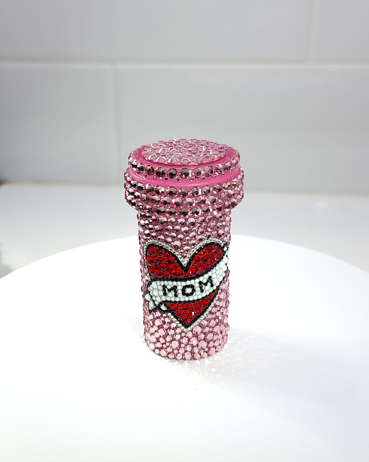 The "MOM" Pill Bottle, a fully rhinestoned pill bottle featuring a heart and banner with the word "mom" written on it, against a pink background.