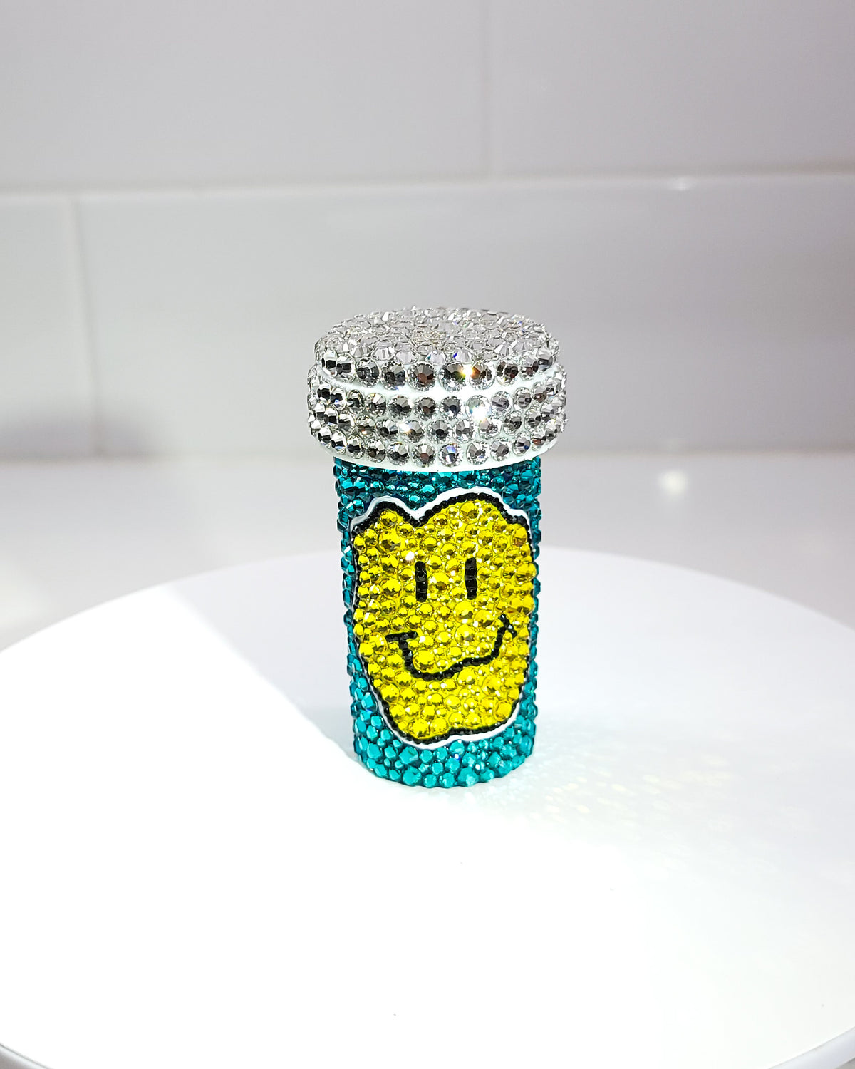 the front of the Smile Harder Pill Bottle, a fully rhinestoned pill bottle featuring a large yellow smiley face against a blue background. 