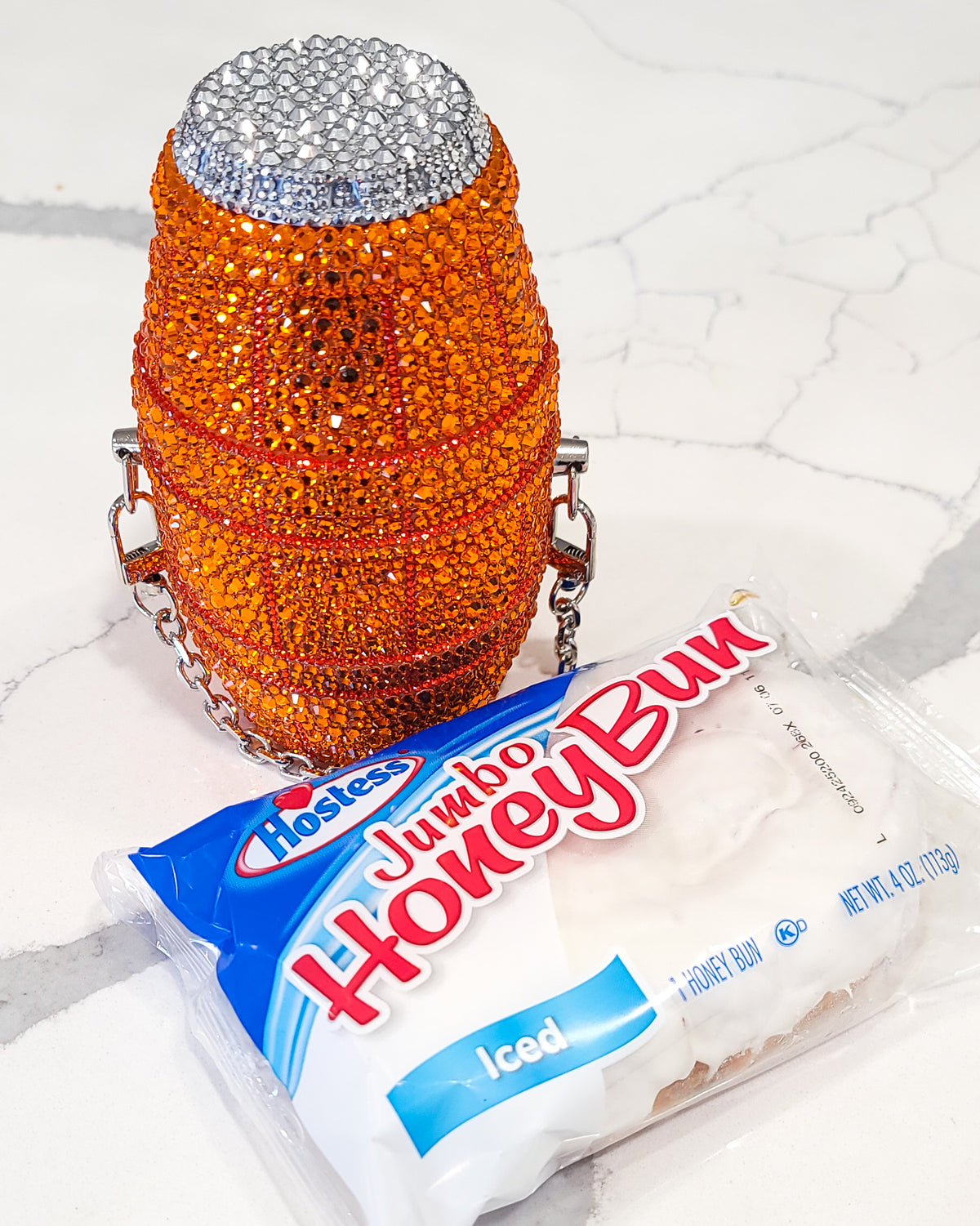 A fully rhinestoned purse, in the shape and style of a quarter water, propped up with a honey bun, against a marble counter top.