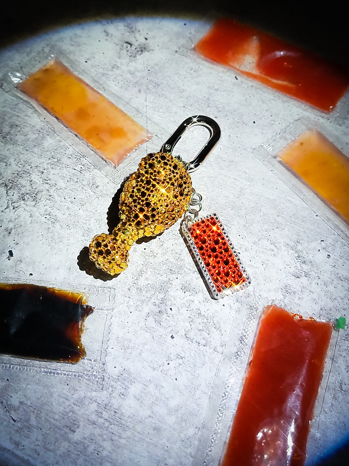 A front view of a chicken drumstick bag charm covered in rhinestones with a tiny hot sauce charm. 