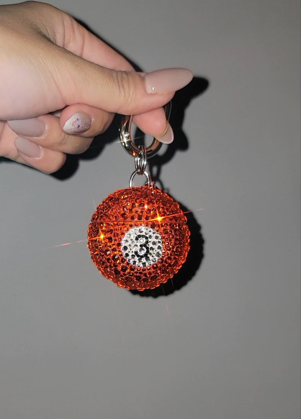 A hand holding the red Pool Ball Bag Charm covered in rhinestones against a gray background