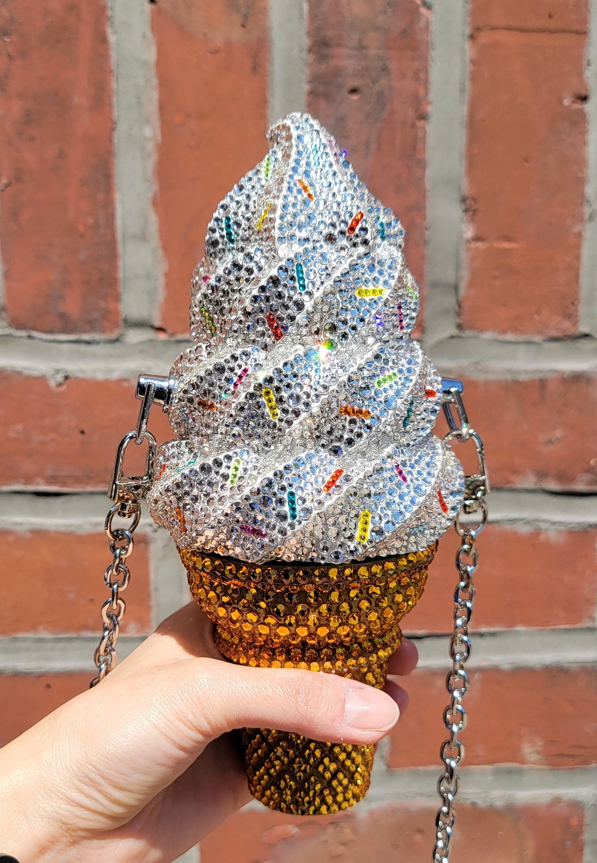A multicolored purse with a rhinestone and crystal embellished design in the shape of an ice cream cone, held by a hand, with a chain strap visible.