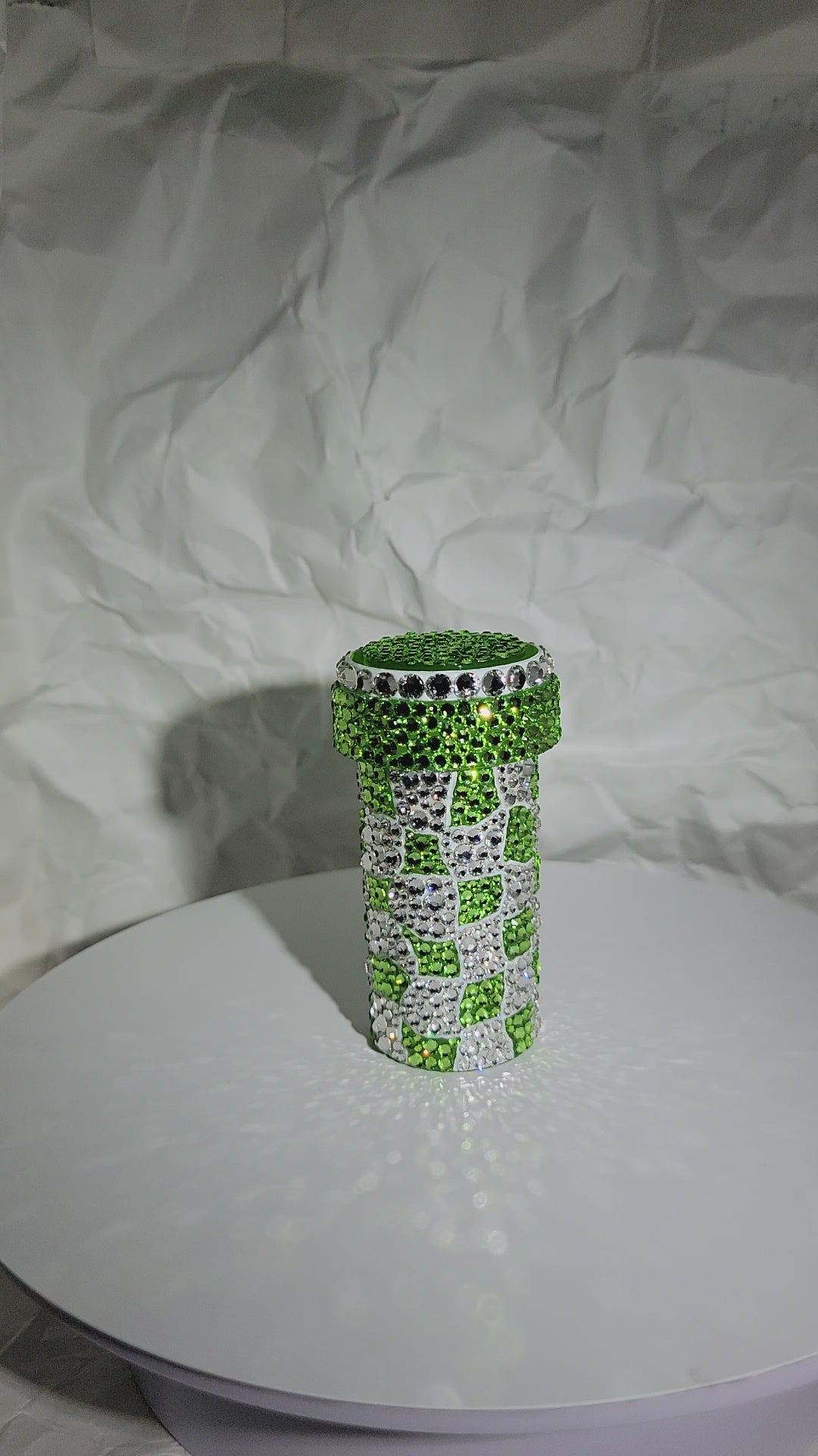 The Trippy Checkerboard Pill Bottle being rotated on a white surface, against a white, crumpled paper background. 
