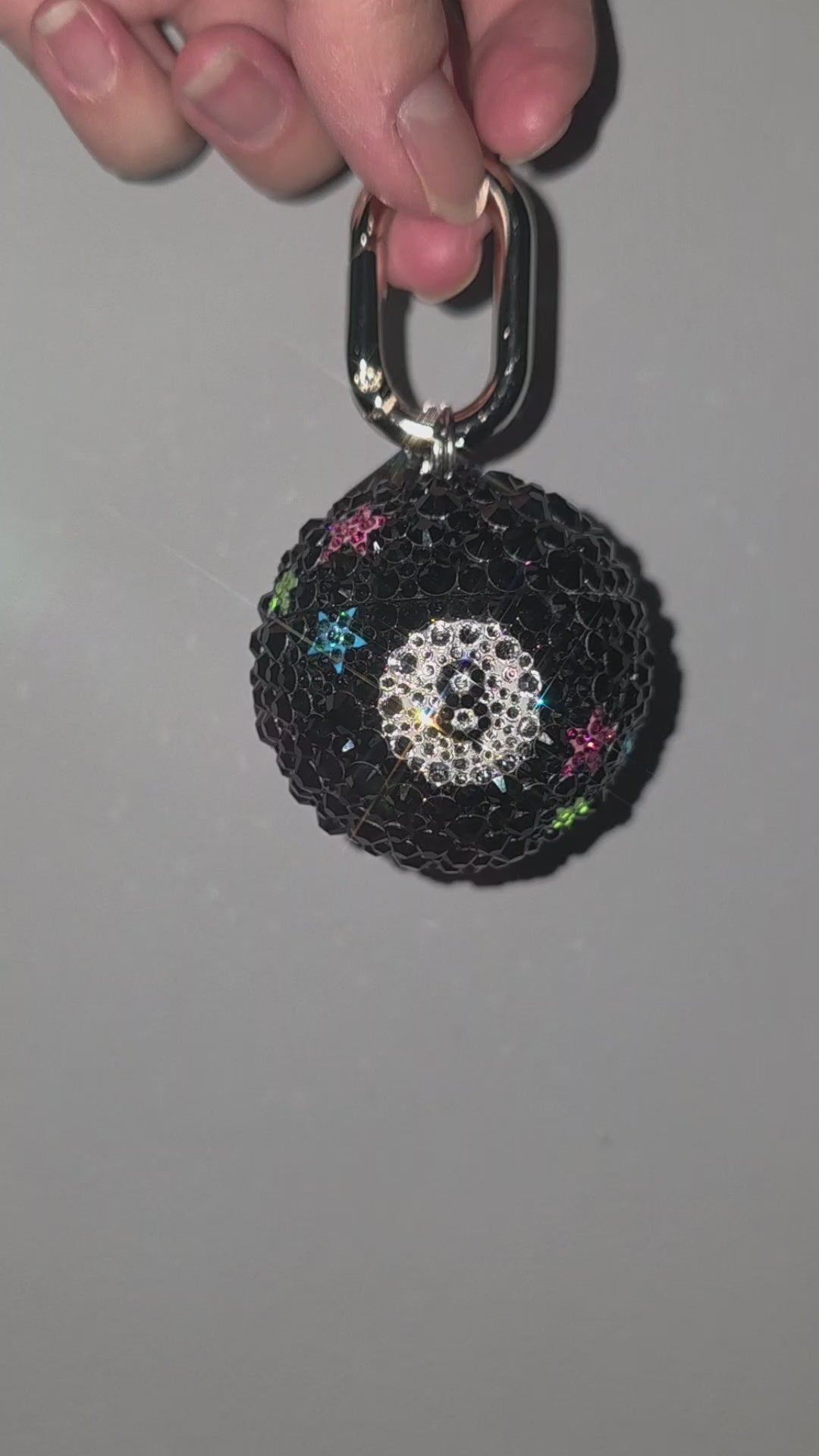 Video of a bag charm resembling an 8 ball covered in rhinestones, being rotated slowly against a gray background.