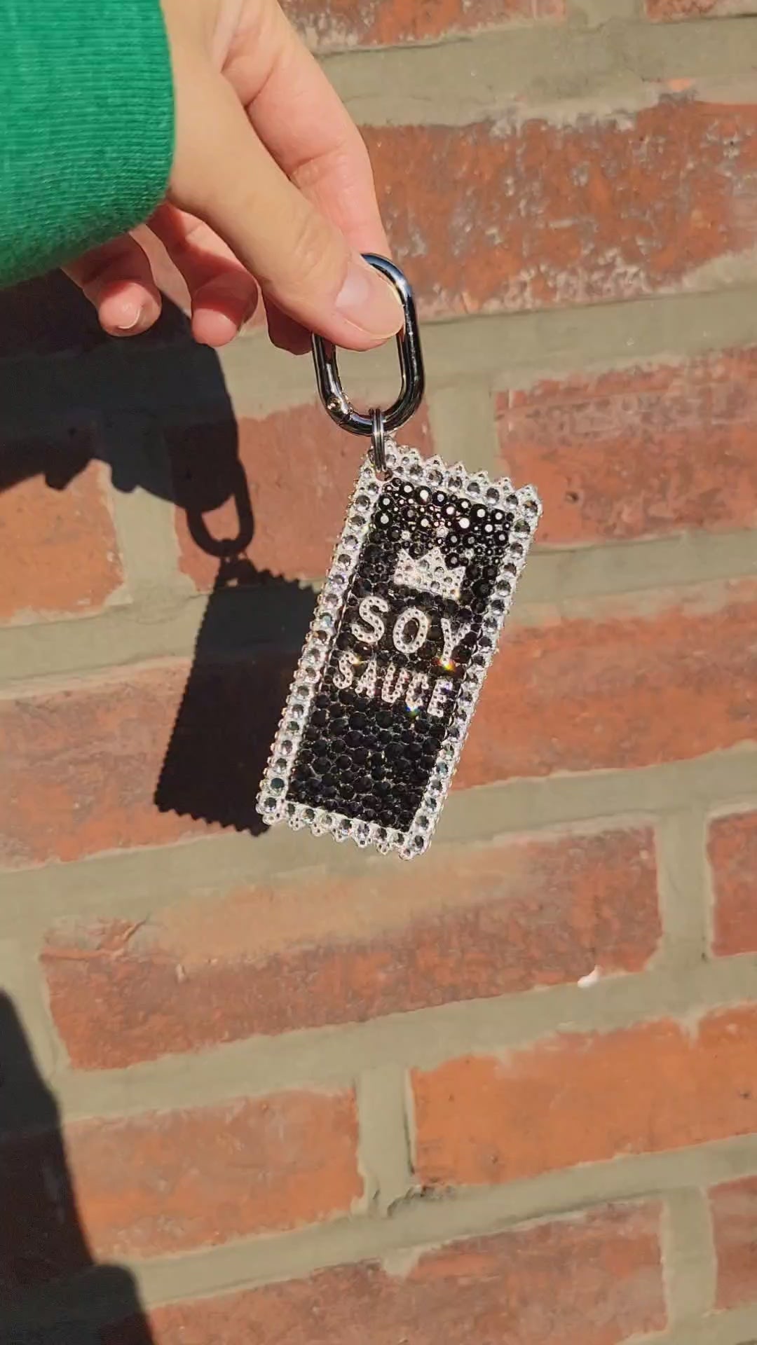 Video of Soy Sauce Bag Charm in the sunlight, held by a hand against a brick background.