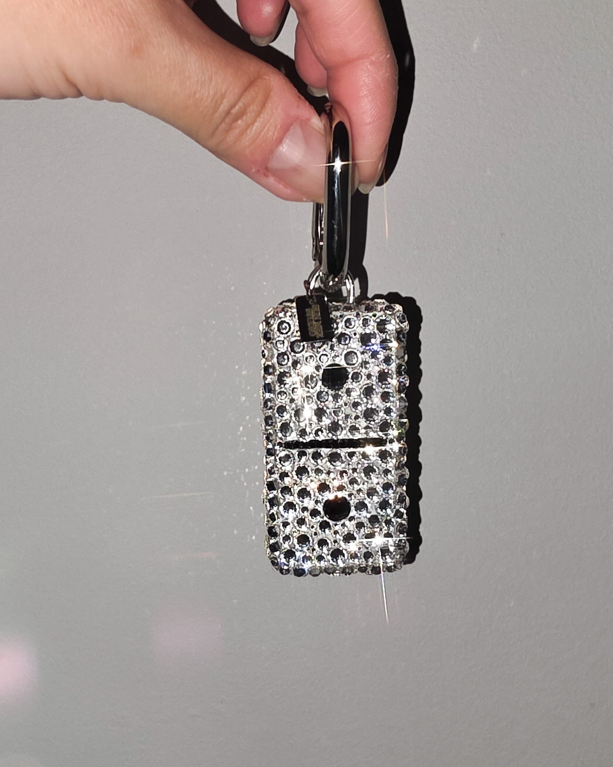Hand holding a 1:1, fully rhinestoned domino bag charm against a gray background.