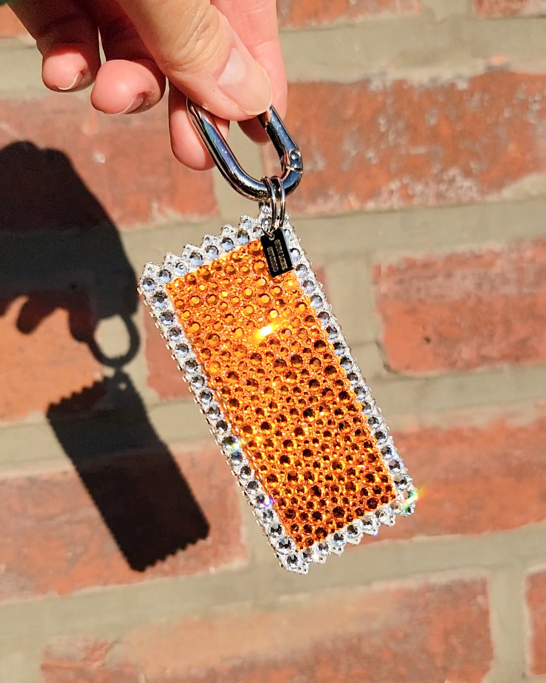 The back of a handmade orange bag charm adorned with Czech Preciosa rhinestones, designed to resemble a duck sauce packet, being held against a brick wall background.
