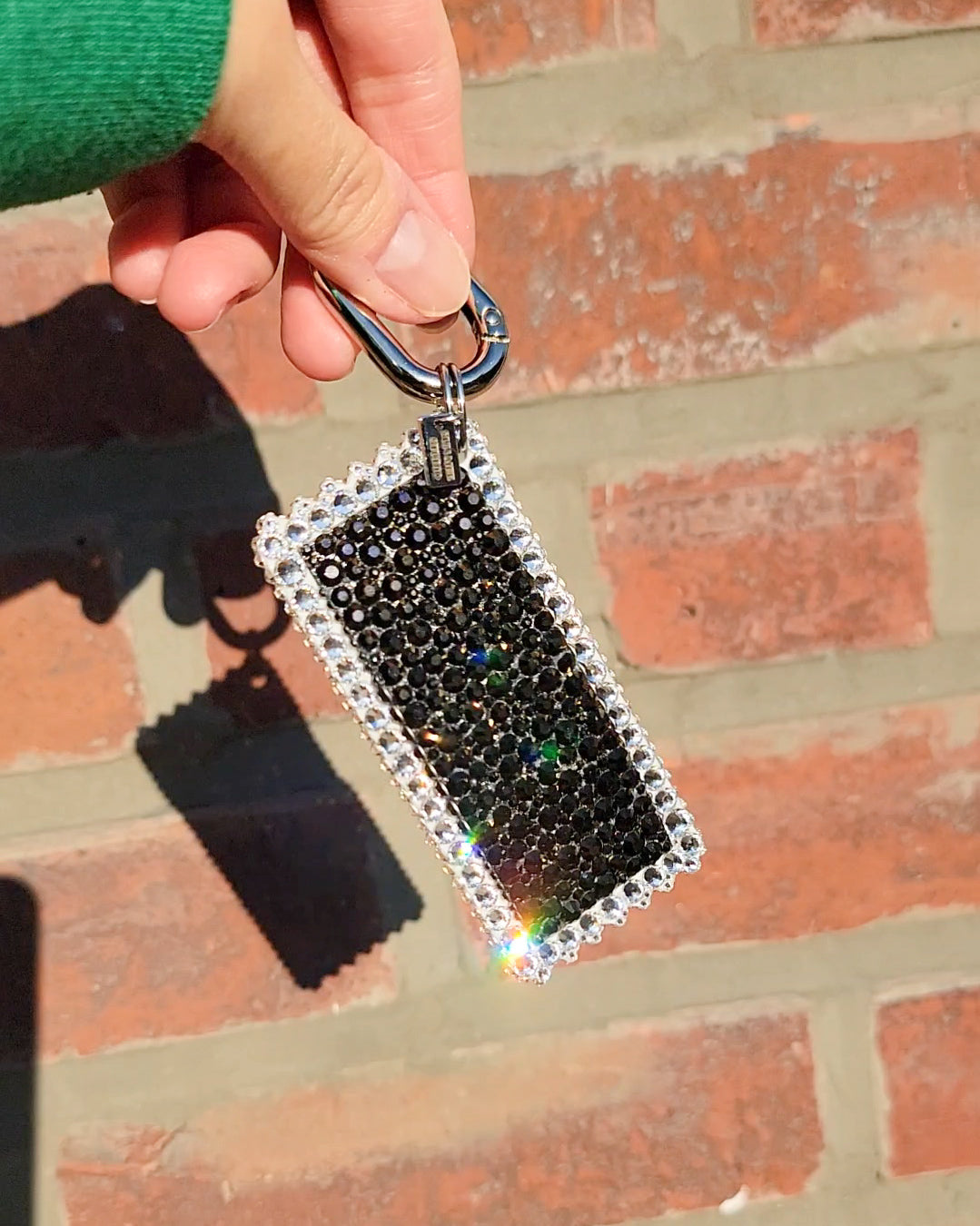 A hand holding a soy sauce bag charm with black background and white text, from the back, adorned with white rhinestones, against a brick wall backdrop.
